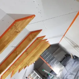 Skośne poddasze w trakcie adaptacji: widoczne drewniane krokwie z pomarańczową taśmą, białe płyty na ścianach i suficie, szare bloczki oraz zabezpieczona folią podłoga.