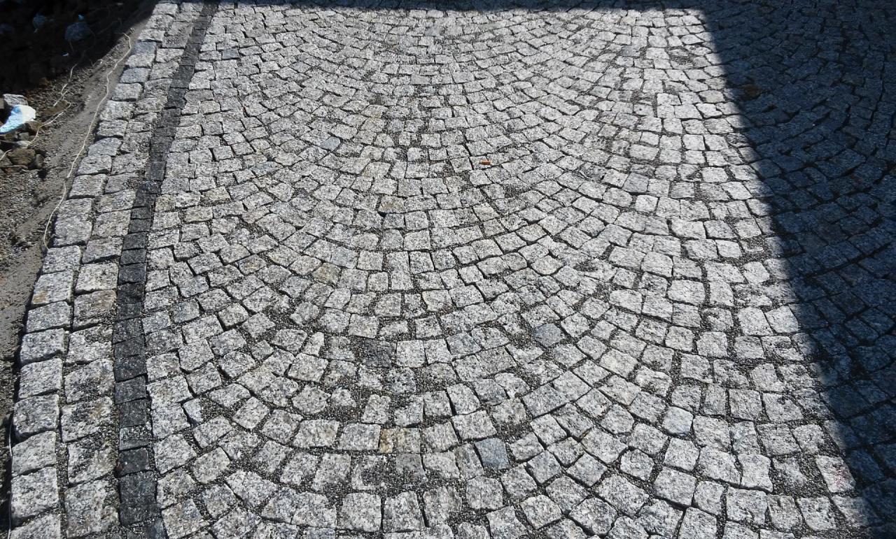 Ułożona z kostki granitowej nawierzchnia w kształcie wachlarza, widoczny fragment obrzeża z ciemniejszej kostki i cień.