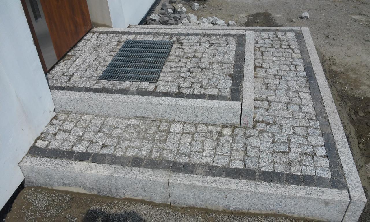 Schody wejściowe wyłożone szarą kostką granitową z ciemną obwódką i kratką ściekową, widoczne resztki piasku po fugowaniu.