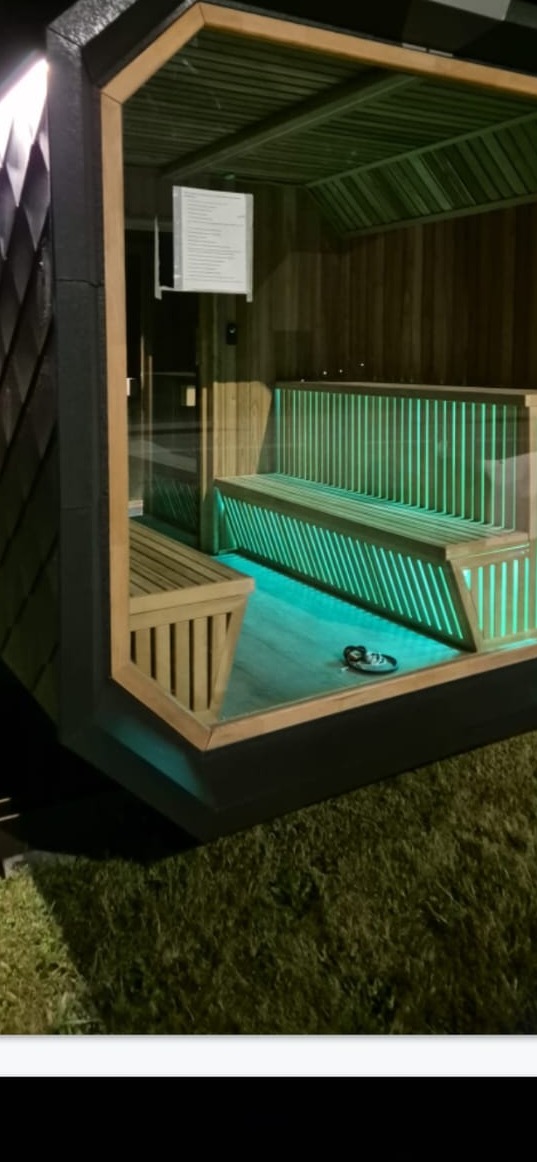 Nowoczesna altana z drewnianymi ławkami podświetlonymi na zielono, czarna konstrukcja zewnętrzna, widok z zewnątrz na tle trawy.