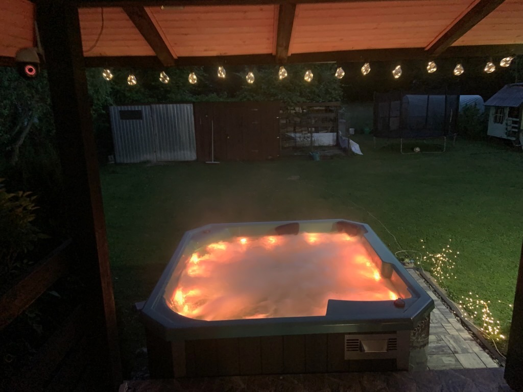 Podświetlane jacuzzi na zewnątrz, otoczone zielonym trawnikiem i dekoracyjnymi lampkami, widok z zadaszonego tarasu. Relaksacyjny klimat.