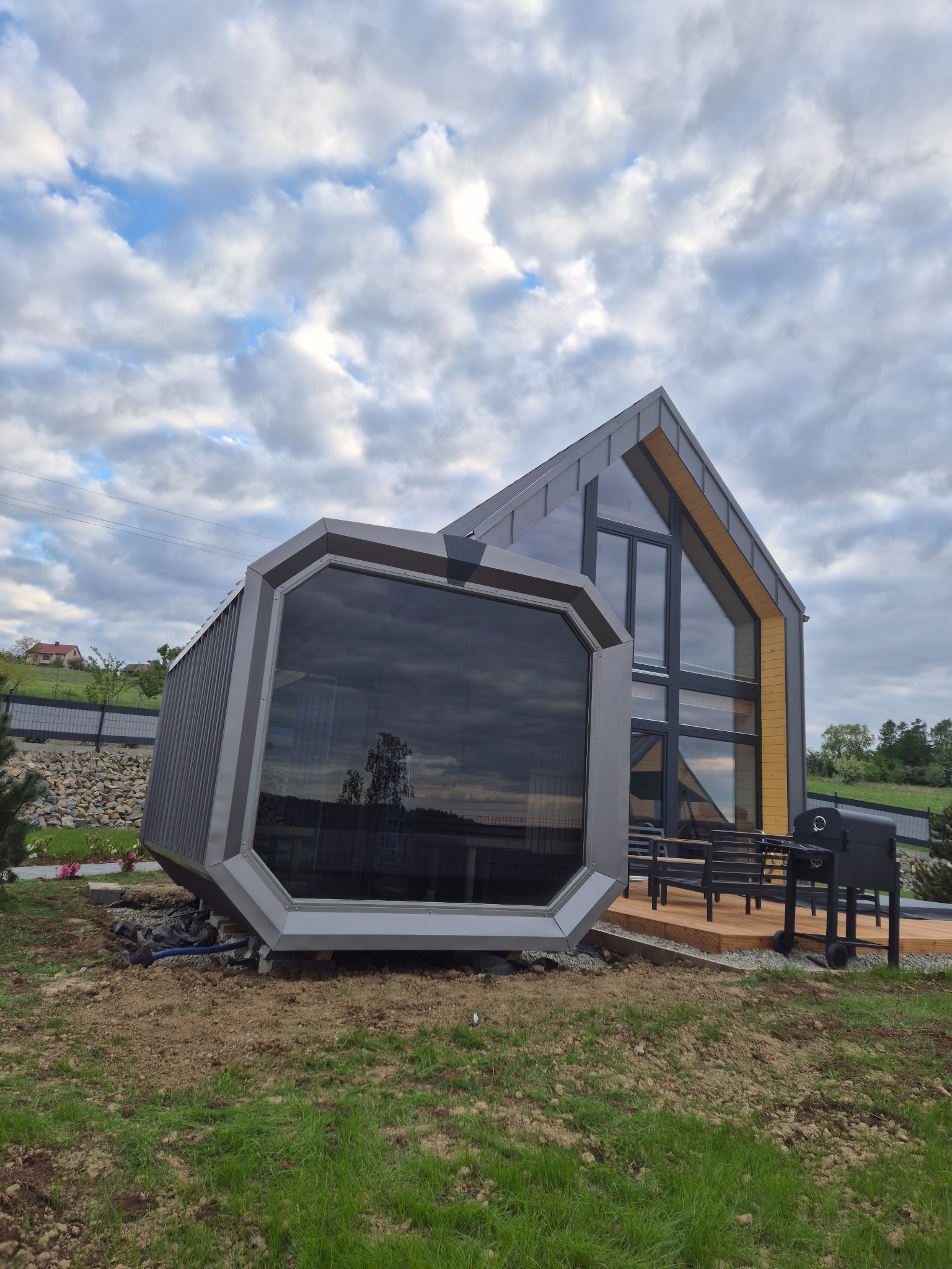 Awangardowy dom z dużymi przeszkleniami i drewnianym tarasem, grill na tarasie, nowoczesna architektura, pochmurne niebo w tle.