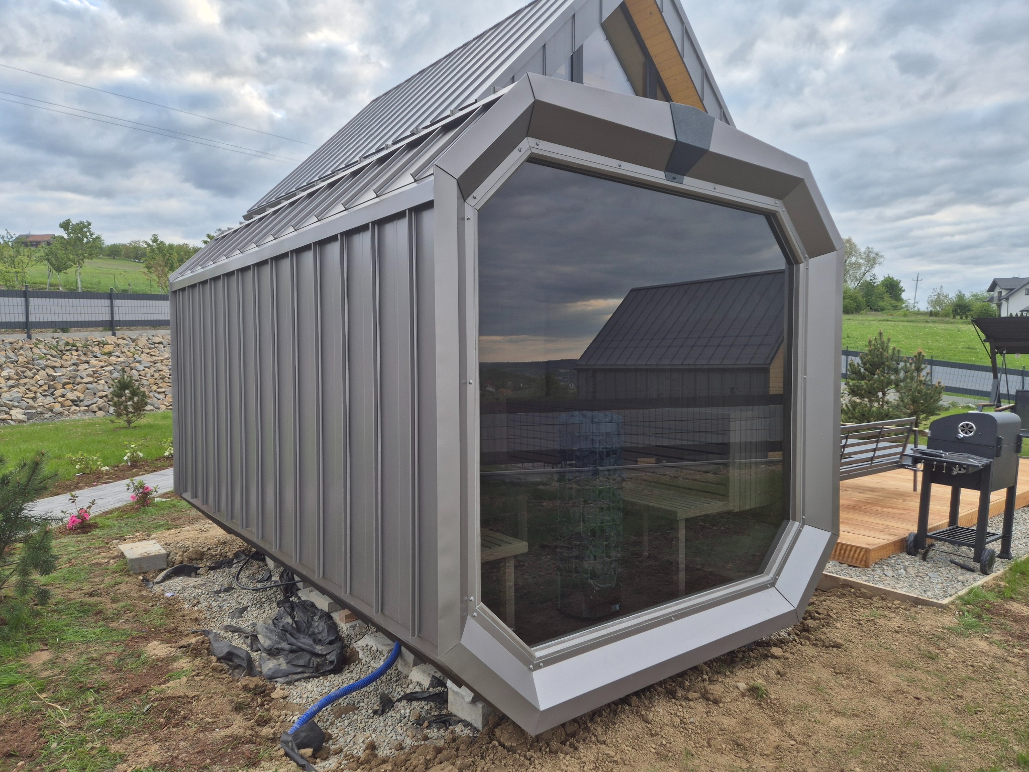 Nowoczesna, metalowa altana z dużym oknem, częściowo osadzona na betonowych blokach. W tle widoczny krajobraz z domami i zielenią. Obok altany grill na drewnianym podeście.