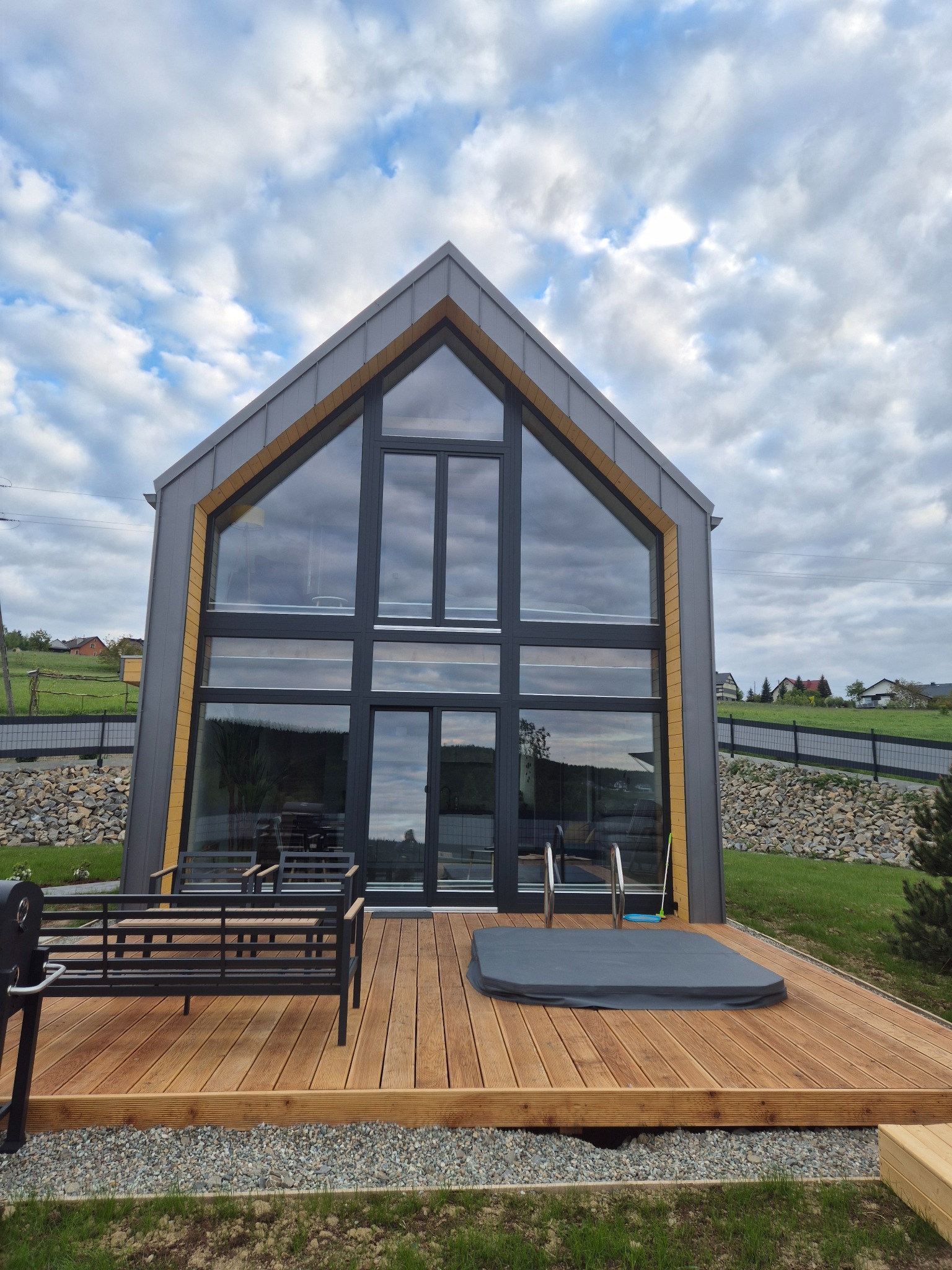 Nowoczesny dom z dużymi oknami i drewnianym tarasem. Na tarasie jacuzzi z pokrywą i metalowa ławka. Tło: zielony trawnik i pochmurne niebo.