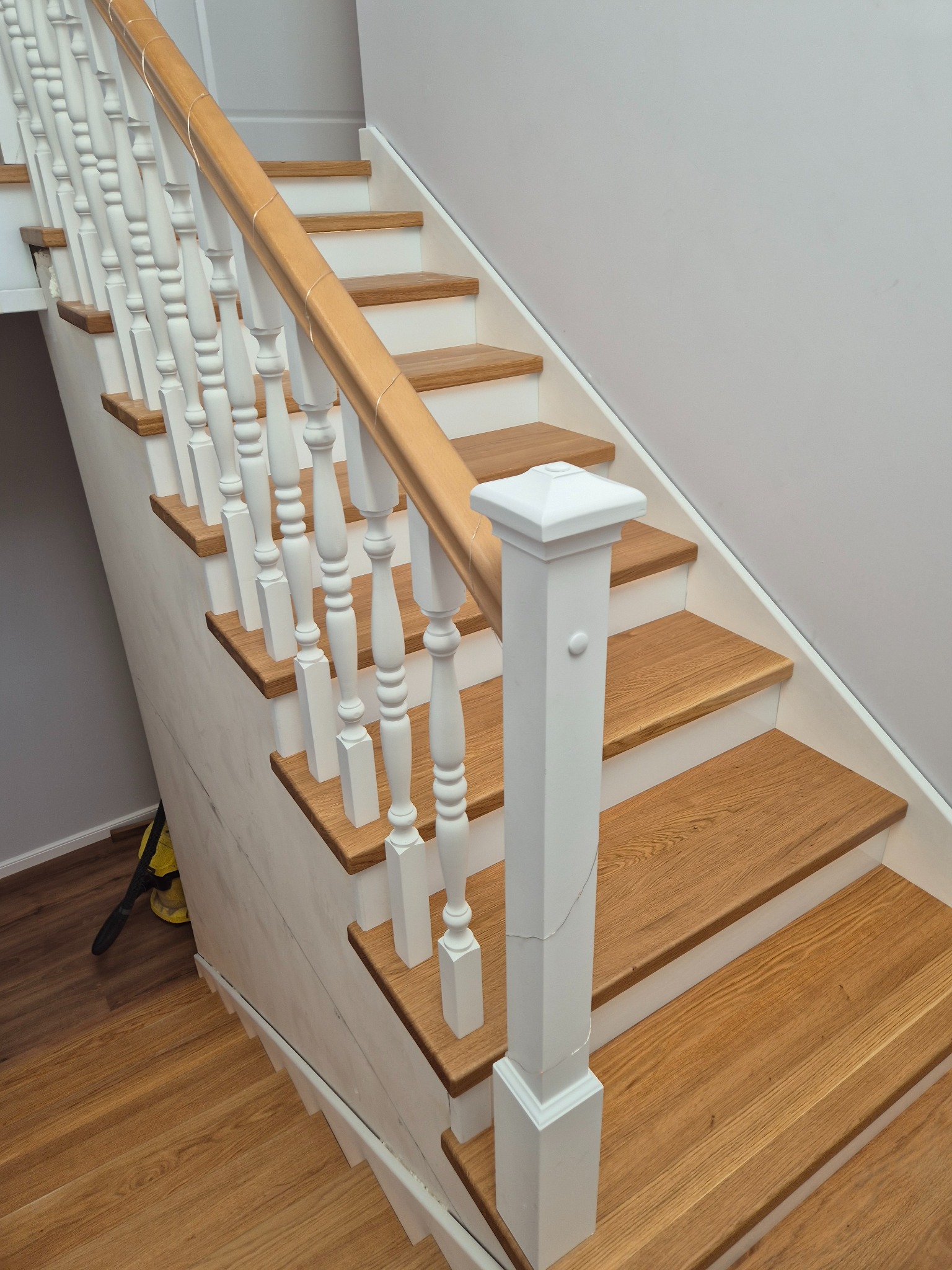 Jasne schody z drewnianymi stopniami i białymi balustradami, widok z dołu. Klasyczny design, naturalne oświetlenie, parkiet na podłodze. Widoczny fragment odkurzacza.
