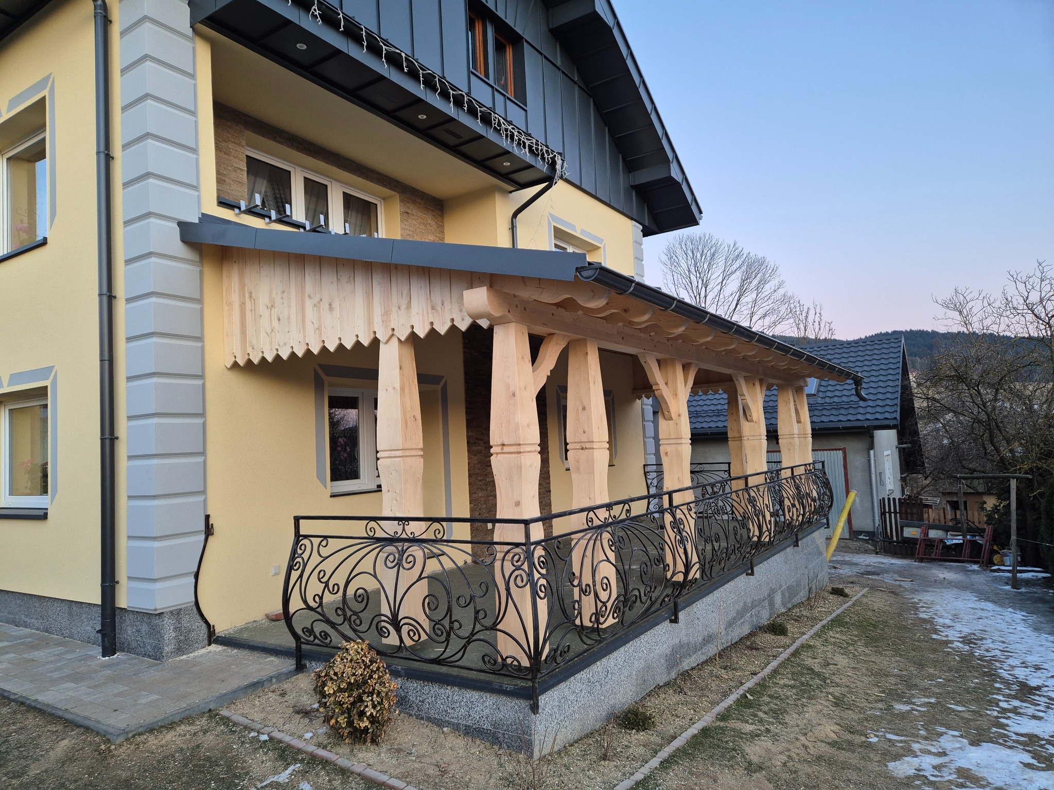 Zadaszony taras z drewnianymi kolumnami i ozdobną, kutą balustradą przy żółtym domu z szarą blachodachówką. Widok z boku, w tle drzewa i fragment sąsiedniego budynku.