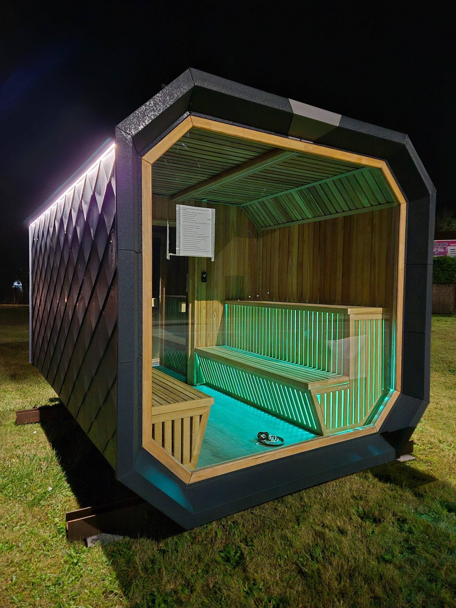Nowoczesna, podświetlana altana z drewnianym wnętrzem i nietypową bryłą, ustawiona na trawie nocą. Widoczne detale wykończenia i oświetlenie LED.