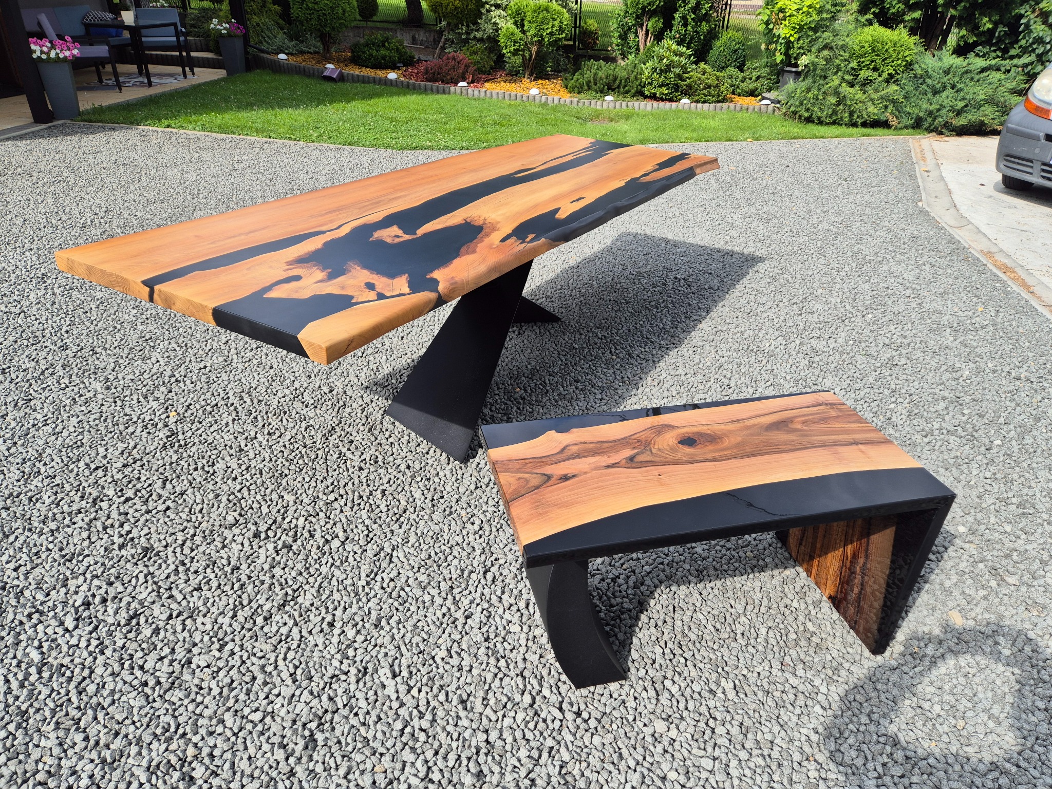 Stół i ława z drewna z żywicą epoksydową na metalowych nogach, ustawione na żwirowym podłożu. Blat stołu z wyraźnym usłojeniem, czarna żywica. Tło: trawnik, krzewy.
