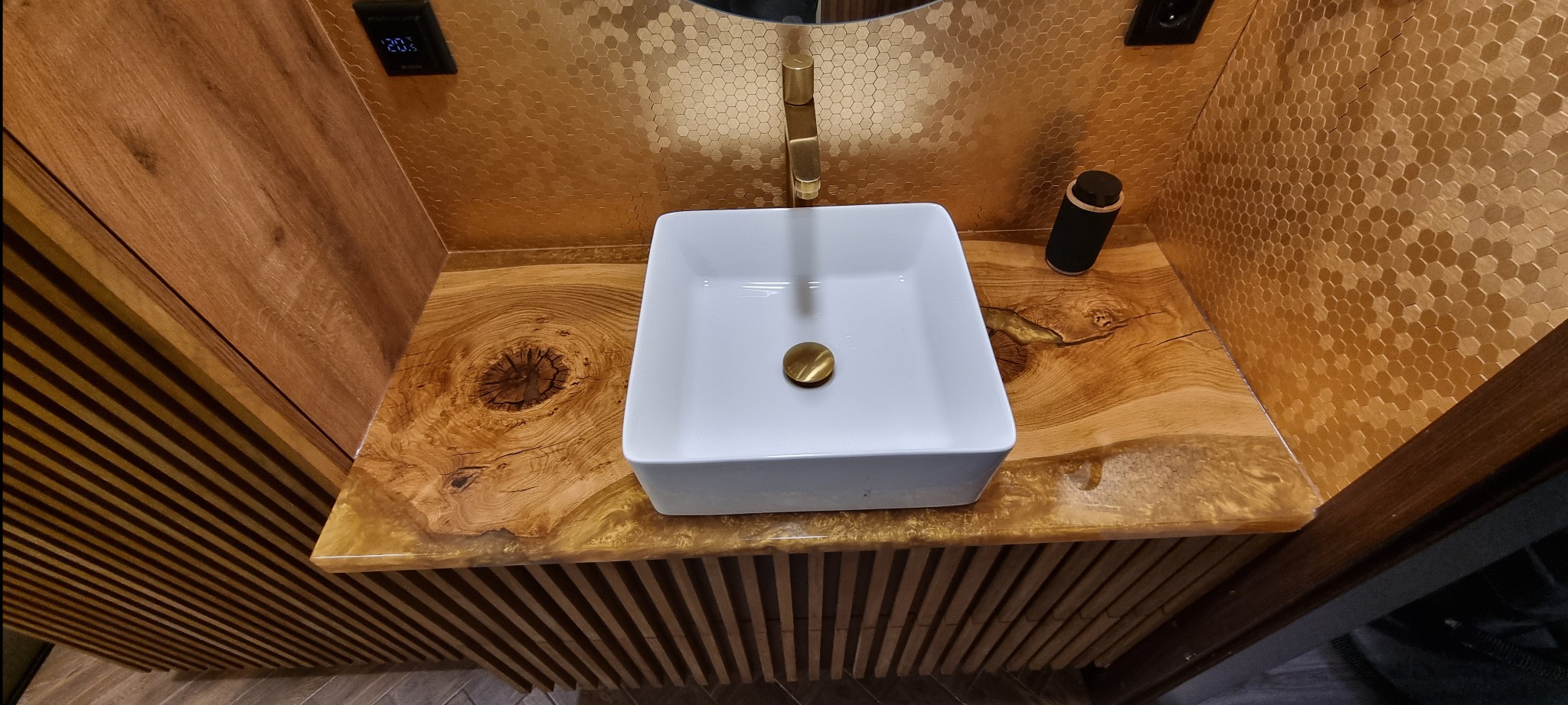 Nowoczesna łazienka z blatem z żywicy epoksydowej i drewna, białą umywalką nablatową i złotą armaturą na tle ściany z mozaiki w kształcie plastra miodu.