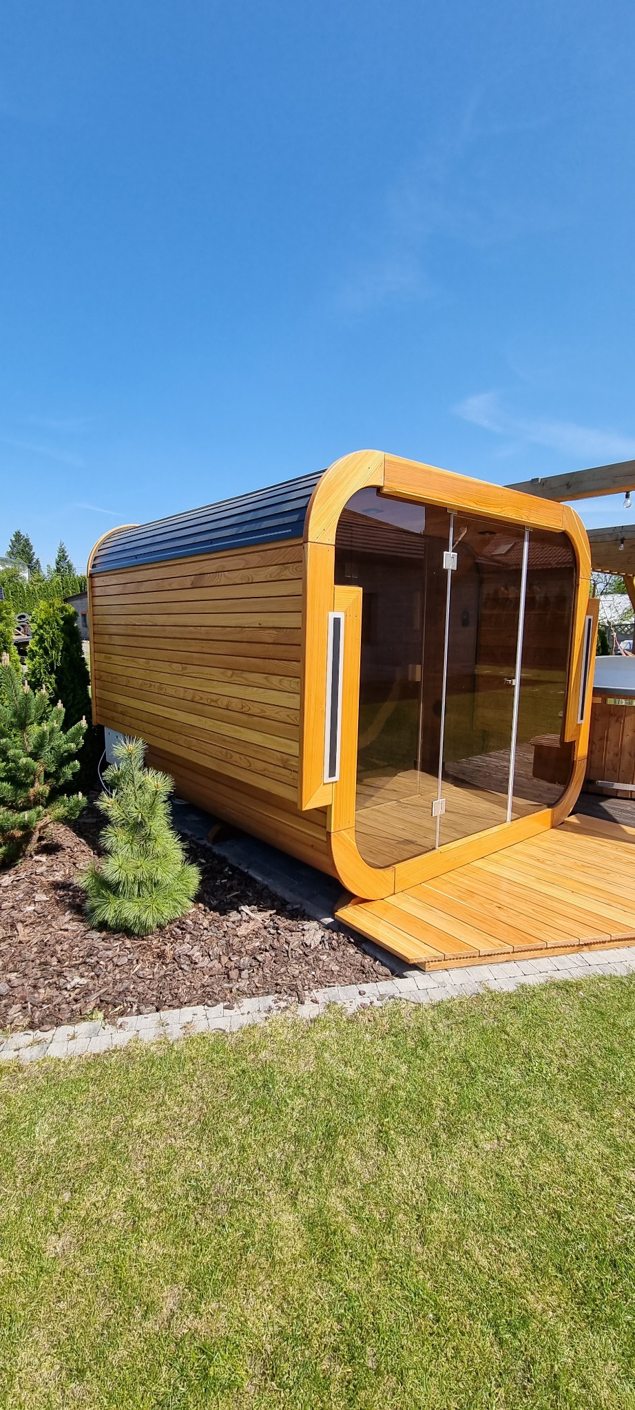 Nowoczesna, zaokrąglona altana z drewna i szkła na trawniku, z widocznym fragmentem tarasu i pergoli w tle, w słoneczny dzień. Unikalny design i naturalne materiały.