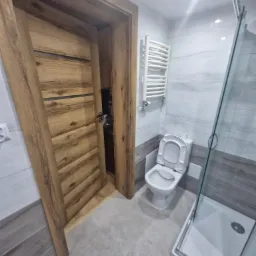 Wykończona łazienka z otwartymi drzwiami z okleiną drewnopodobną, widoczna toaleta z otwartą klapą, grzejnik drabinkowy i szklana kabina prysznicowa.