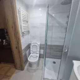 Wykończona łazienka w Działdowie z kabiną prysznicową typu walk-in, podwieszaną toaletą z otwartą klapą i grzejnikiem drabinkowym na ścianie wyłożonej płytkami imitującymi marmur.