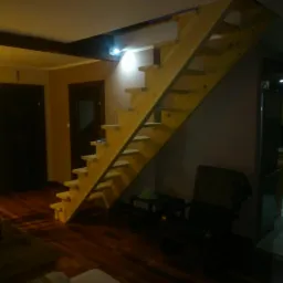 Drewniane schody bez poręczy, z widoczną konstrukcją stopni, wkomponowane w przestrzeń mieszkalną z drewnianą podłogą i widoczną kuchnią w tle.