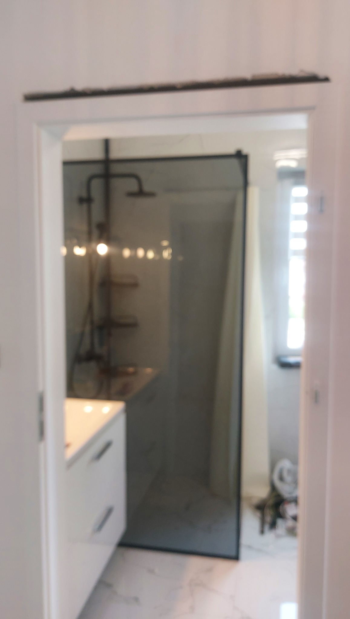 Nowoczesna łazienka z kabiną prysznicową typu walk-in z czarnymi profilami, białą szafką z umywalką i marmurową posadzką, widok zza drzwi.