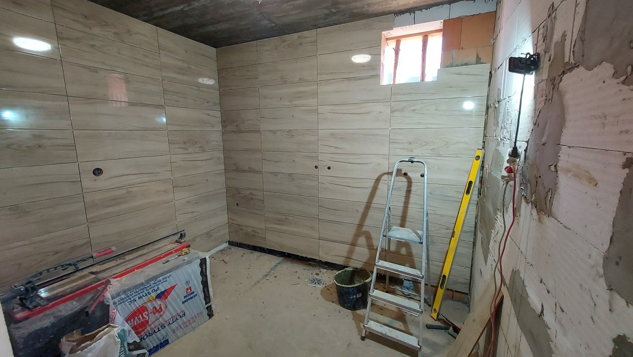 Układanie płytek imitujących drewno w trakcie remontu łazienki, widoczna drabina, poziomica, narzędzia glazurnicze i materiały budowlane, surowy betonowy sufit i fragment ściany bez płytek...