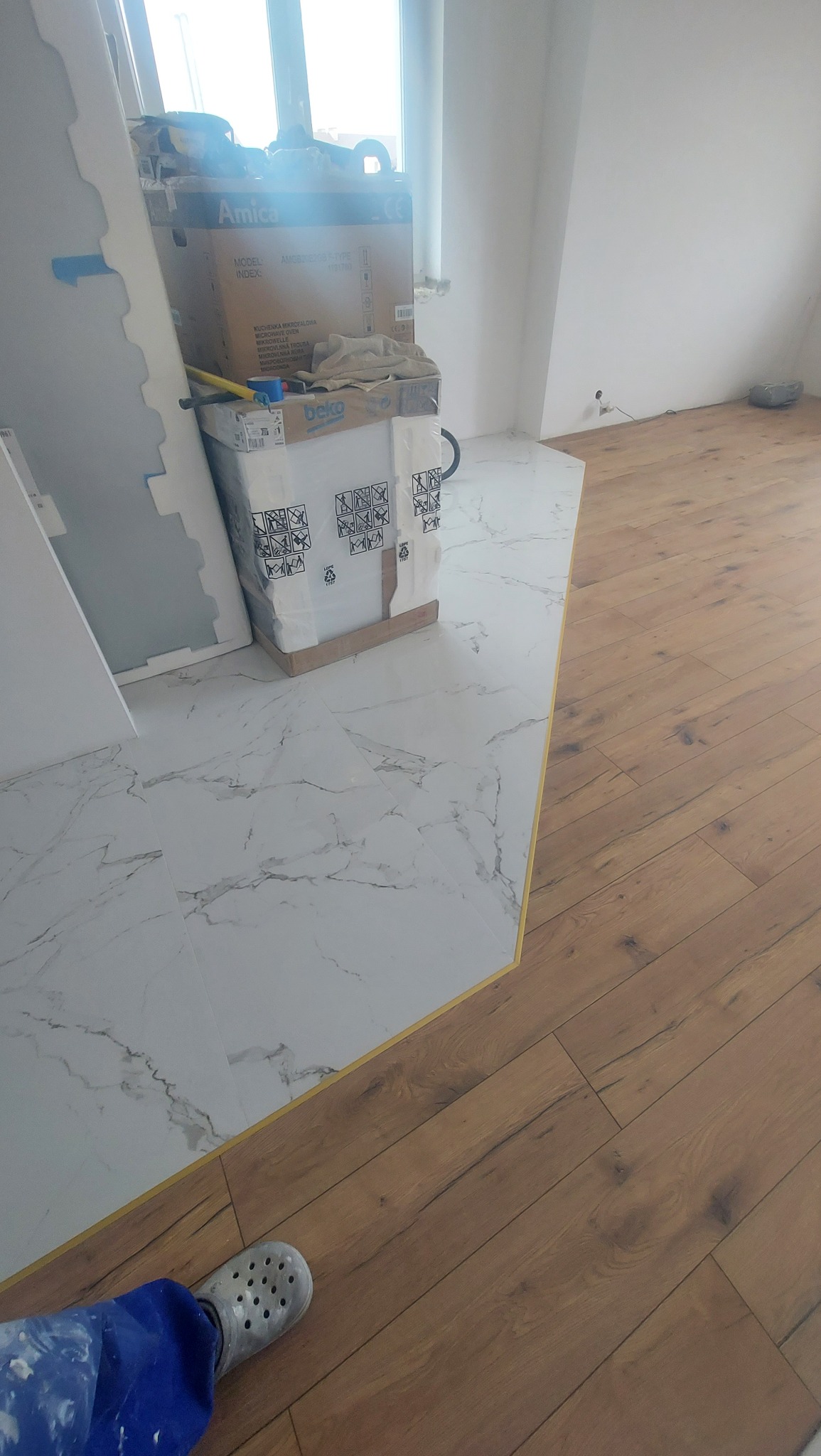Połączenie podłogi z płytek imitujących marmur i paneli drewnopodobnych, widoczna listwa przejściowa, kartony i narzędzia w tle, fragment roboczego ubrania i buta.