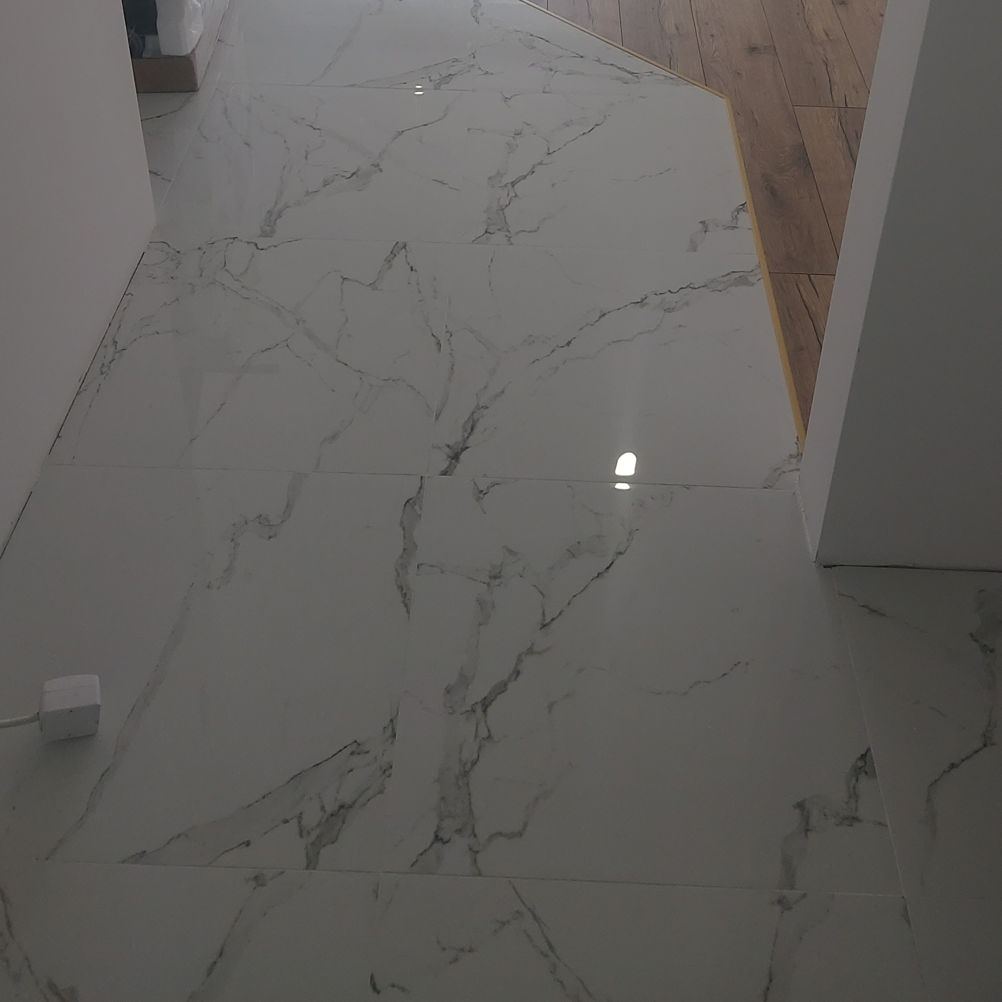 Białe płytki podłogowe imitujące marmur z widocznymi fugami, przechodzące w drewnianą podłogę, z białym kablem zasilającym w lewym dolnym rogu.