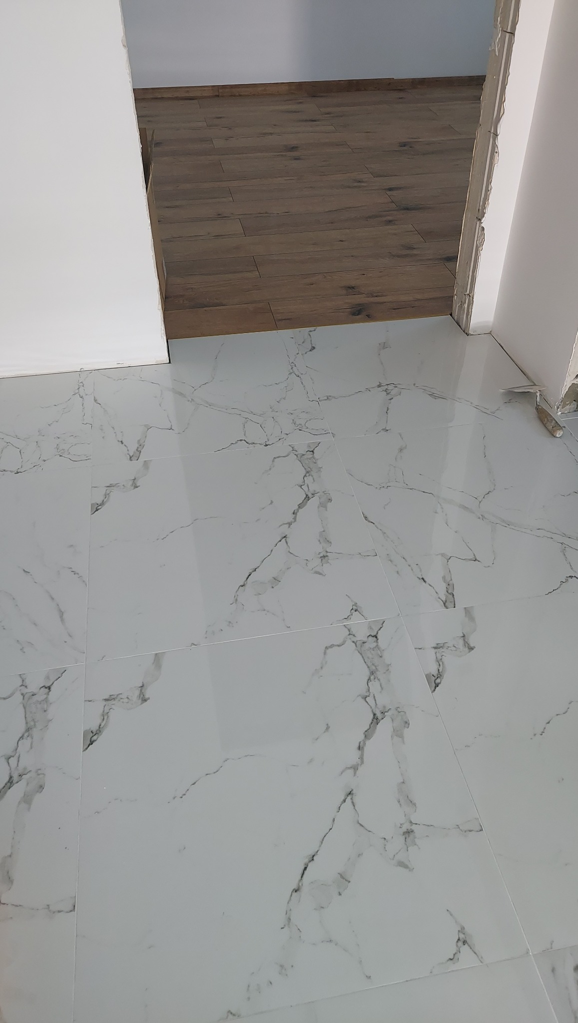Białe płytki podłogowe z marmurkowym wzorem, świeżo ułożone, widoczna fuga, częściowo widoczne drewniane panele w sąsiednim pomieszczeniu, narzędzie do fugowania w rogu.
