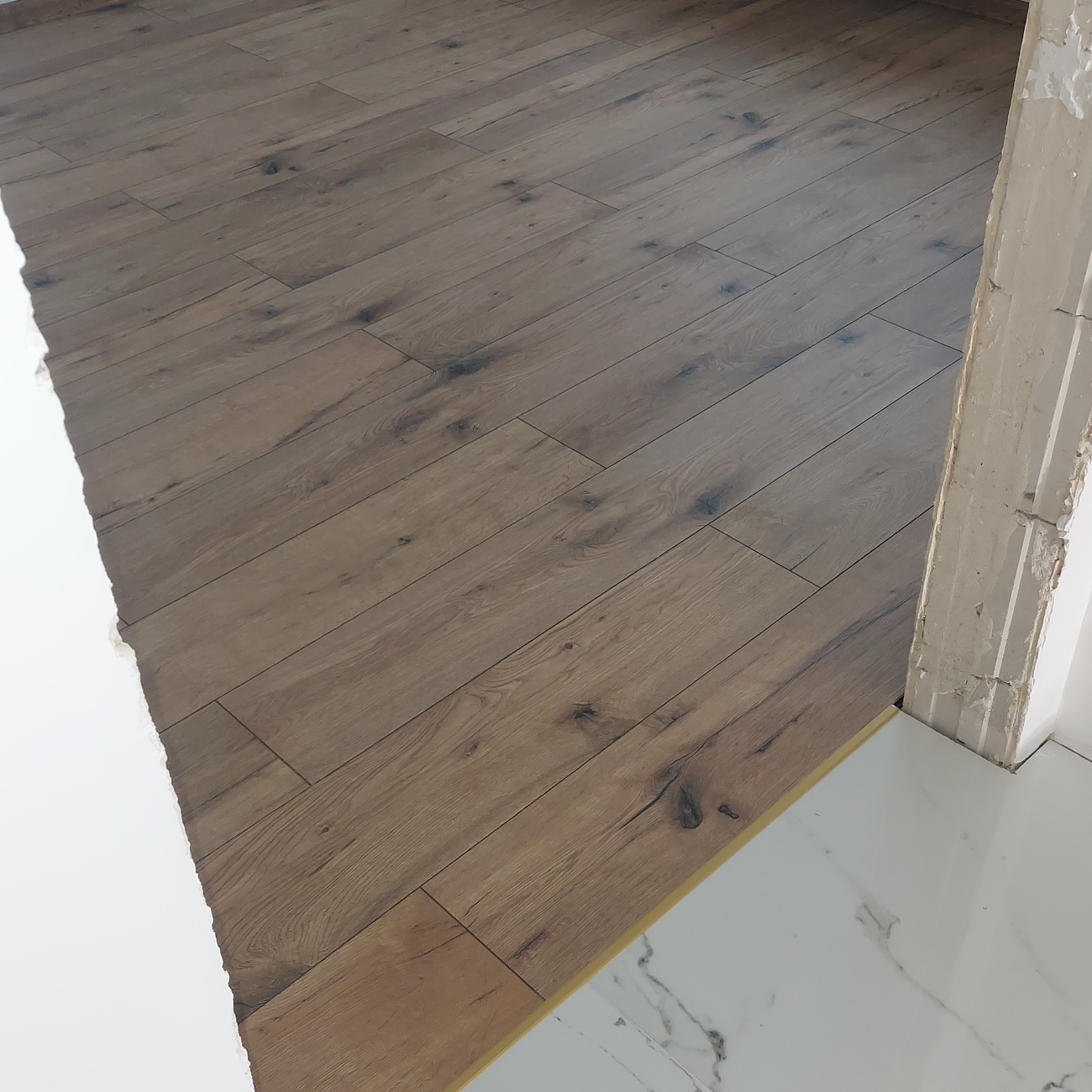 Ułożona podłoga z paneli imitujących drewno, widoczna listwa przypodłogowa oraz fragment wykończonej białej podłogi z płytek imitujących marmur, krawędź ściany w trakcie wykończenia.