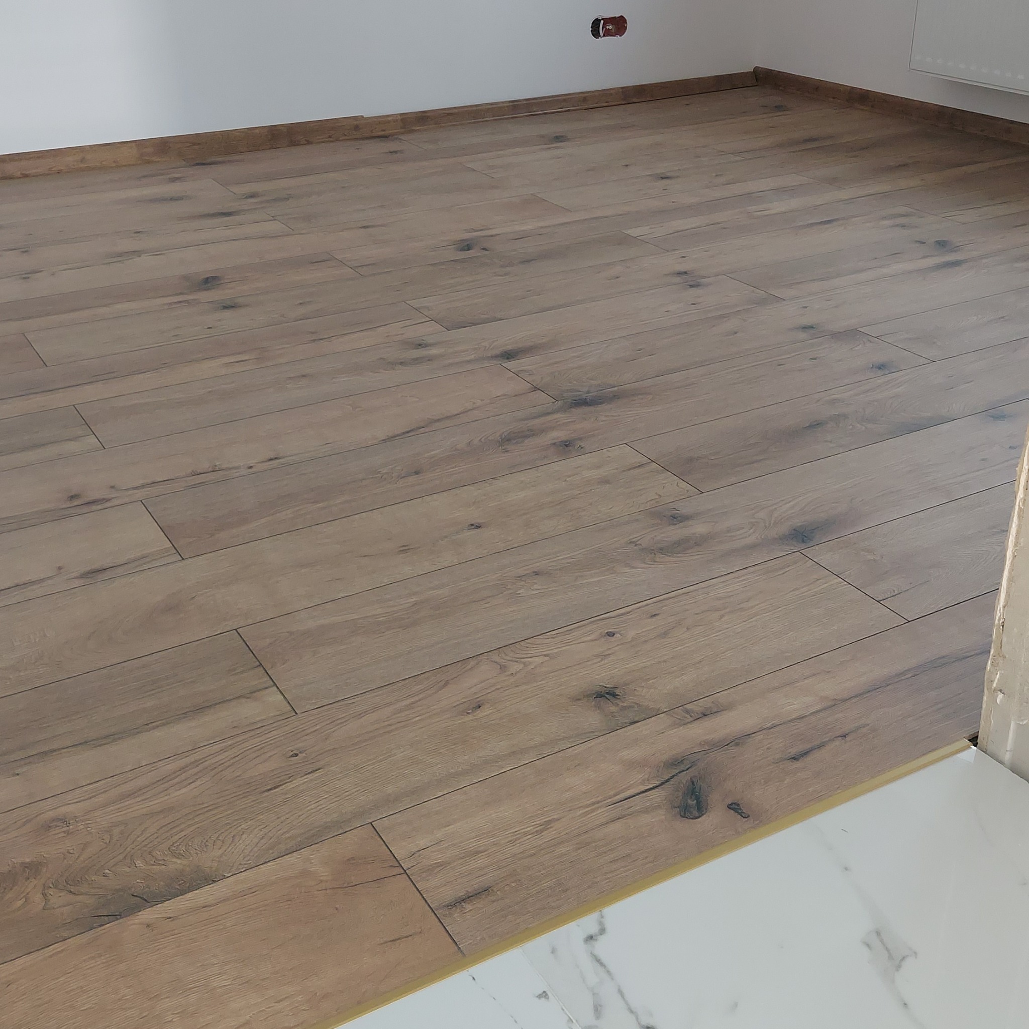 Ułożona podłoga z paneli imitujących drewno, widoczne listwy przypodłogowe i fragment białej marmurowej posadzki w sąsiednim pomieszczeniu, widoczny też niedokończony fragment ściany.