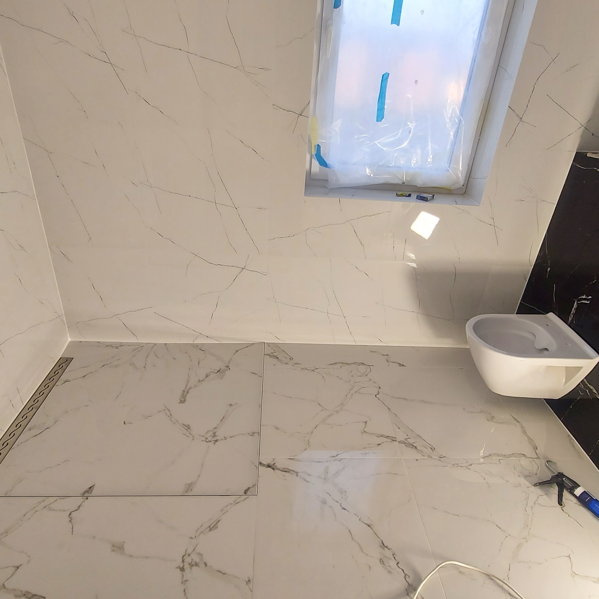 Białe płytki imitujące marmur na podłodze i ścianach łazienki, widoczny odpływ liniowy, okno dachowe zabezpieczone folią, częściowo zamontowana toaleta wisząca i pistolet do silikonu.