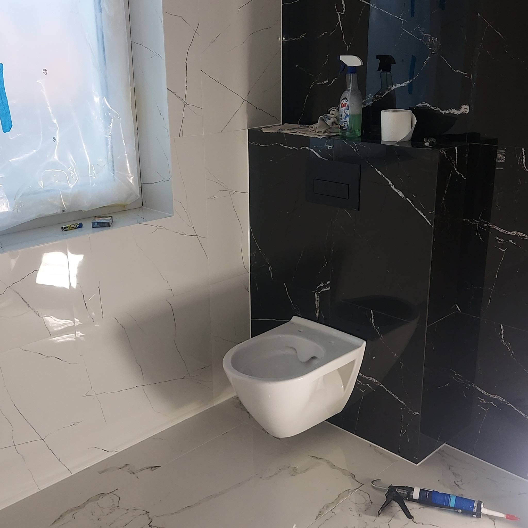 Montaż podwieszanej toalety na tle czarno-białych płytek imitujących marmur, widoczne narzędzia glazurnicze i środki czystości.