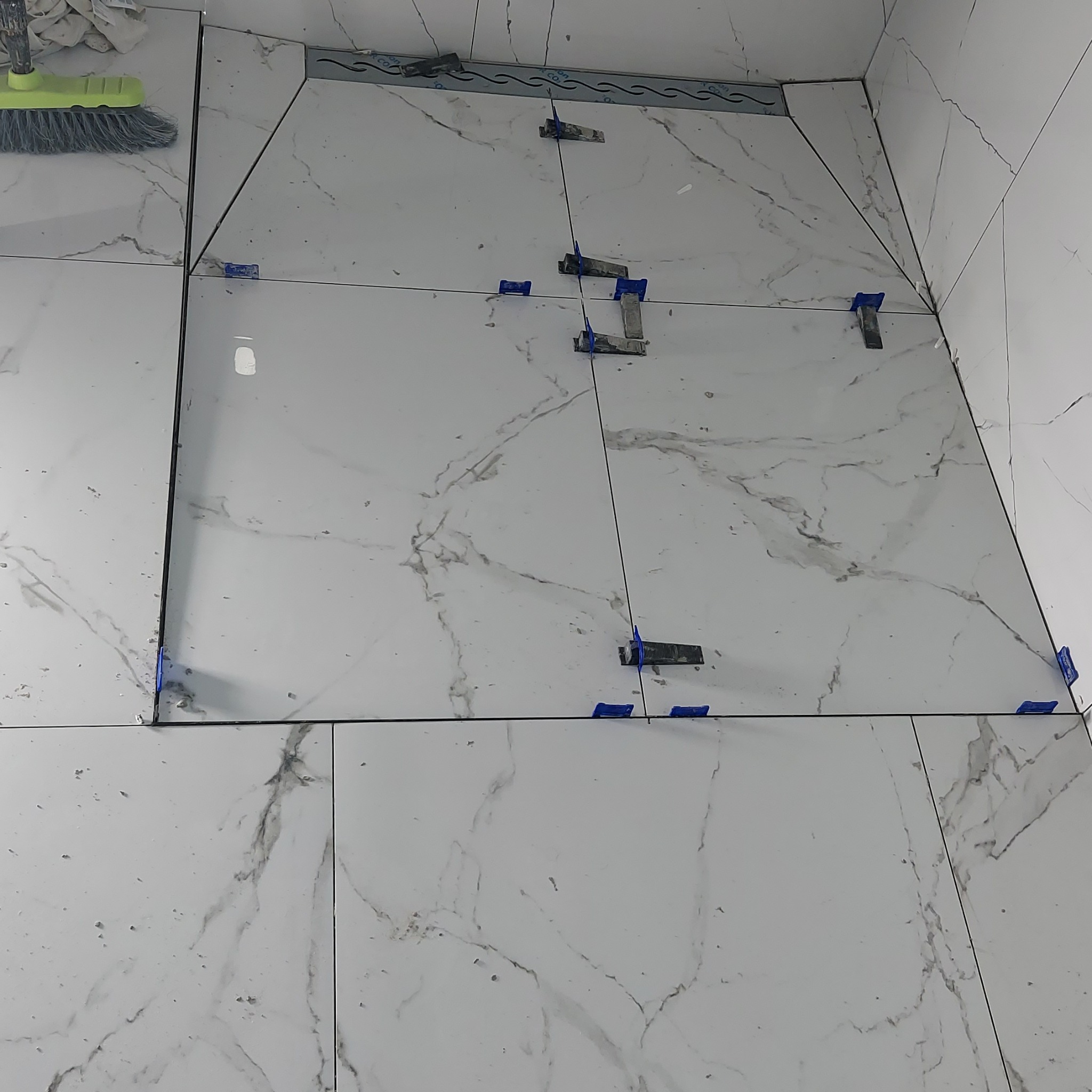 Białe płytki imitujące marmur, ułożone na podłodze i ścianie, z użyciem niebieskich klipsów poziomujących i listwy przypodłogowej z odpływem liniowym.
