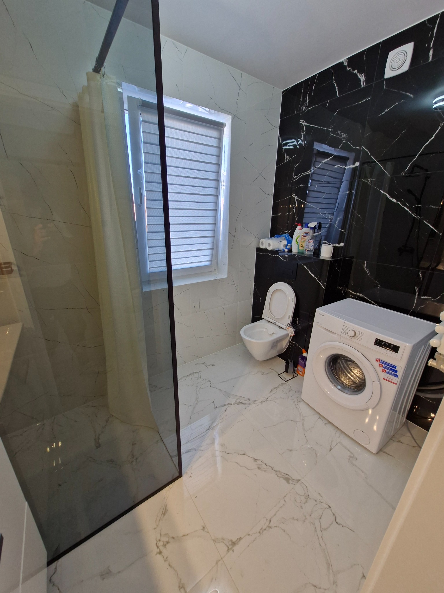 Nowoczesna łazienka z marmurowymi płytkami, kabiną prysznicową typu walk-in z czarną ramą, białą toaletą, pralką i akcesoriami na półce.