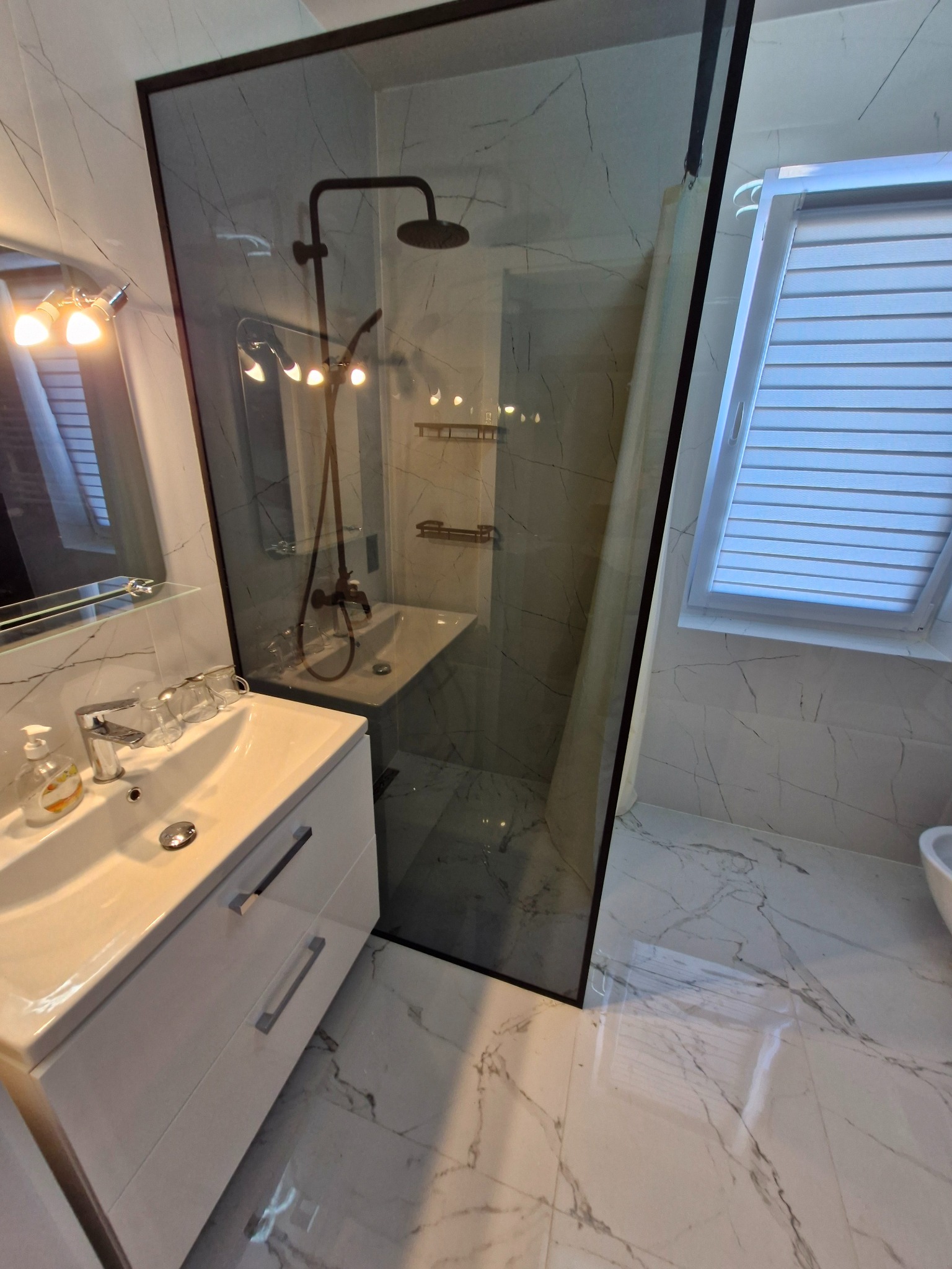 Nowoczesna łazienka z kabiną prysznicową typu walk-in z czarną ramą, umywalką z szafką w kolorze białym, armaturą w kolorze czarnym i marmurową okładziną ścian i podłogi.