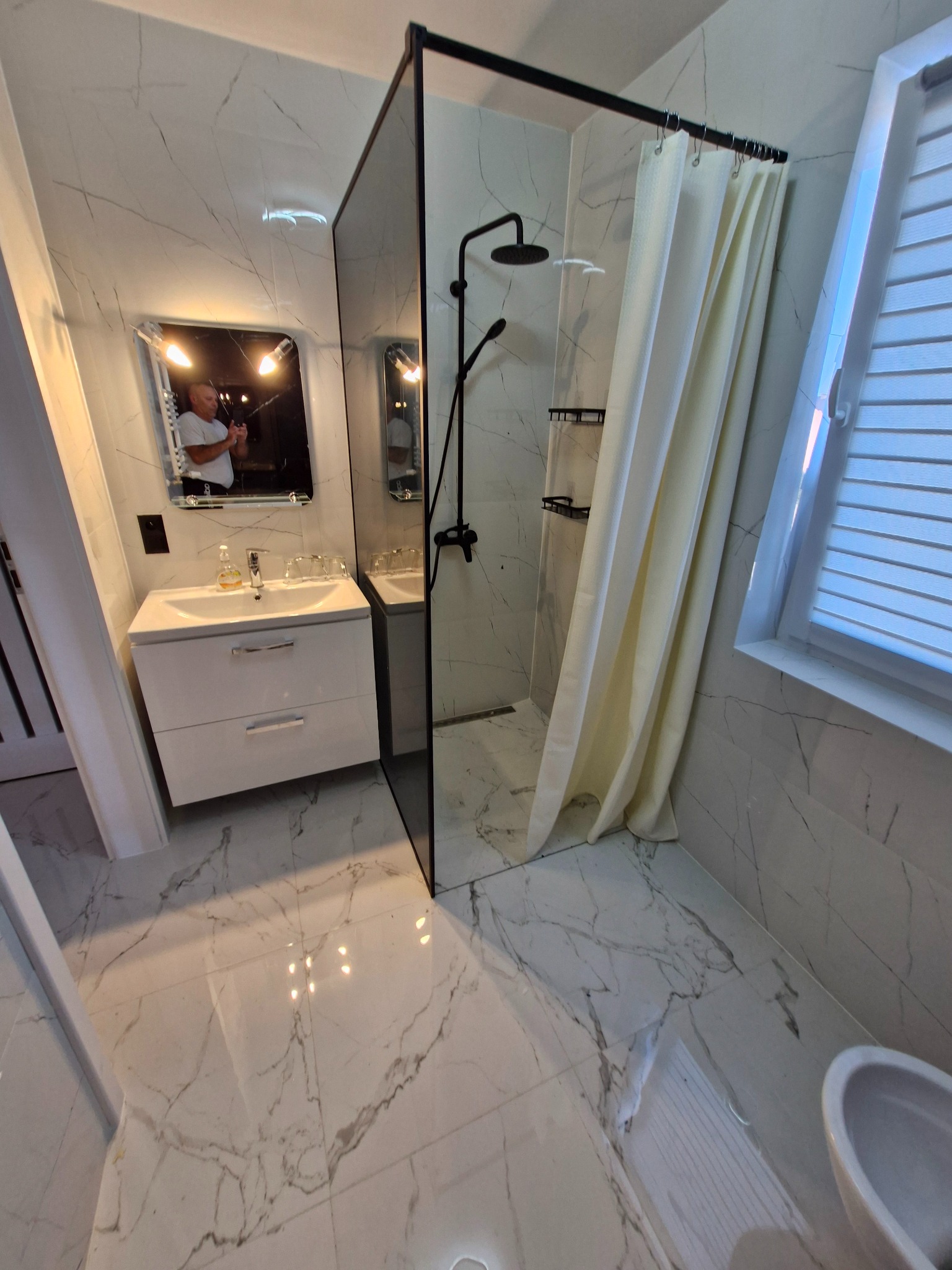 Wykończona łazienka z kabiną prysznicową typu walk-in, białymi marmurkowymi płytkami na ścianach i podłodze, wiszącą szafką z umywalką i lustrem z odbijającą się postacią.