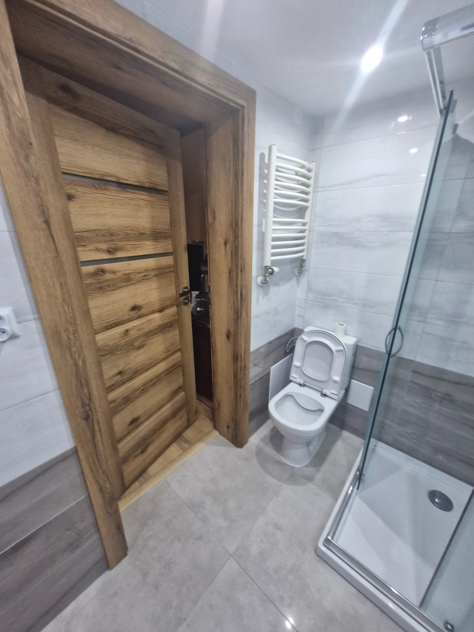 Wykończona łazienka z otwartymi drzwiami z okleiną drewnopodobną, widoczna toaleta z otwartą klapą, grzejnik drabinkowy i szklana kabina prysznicowa.