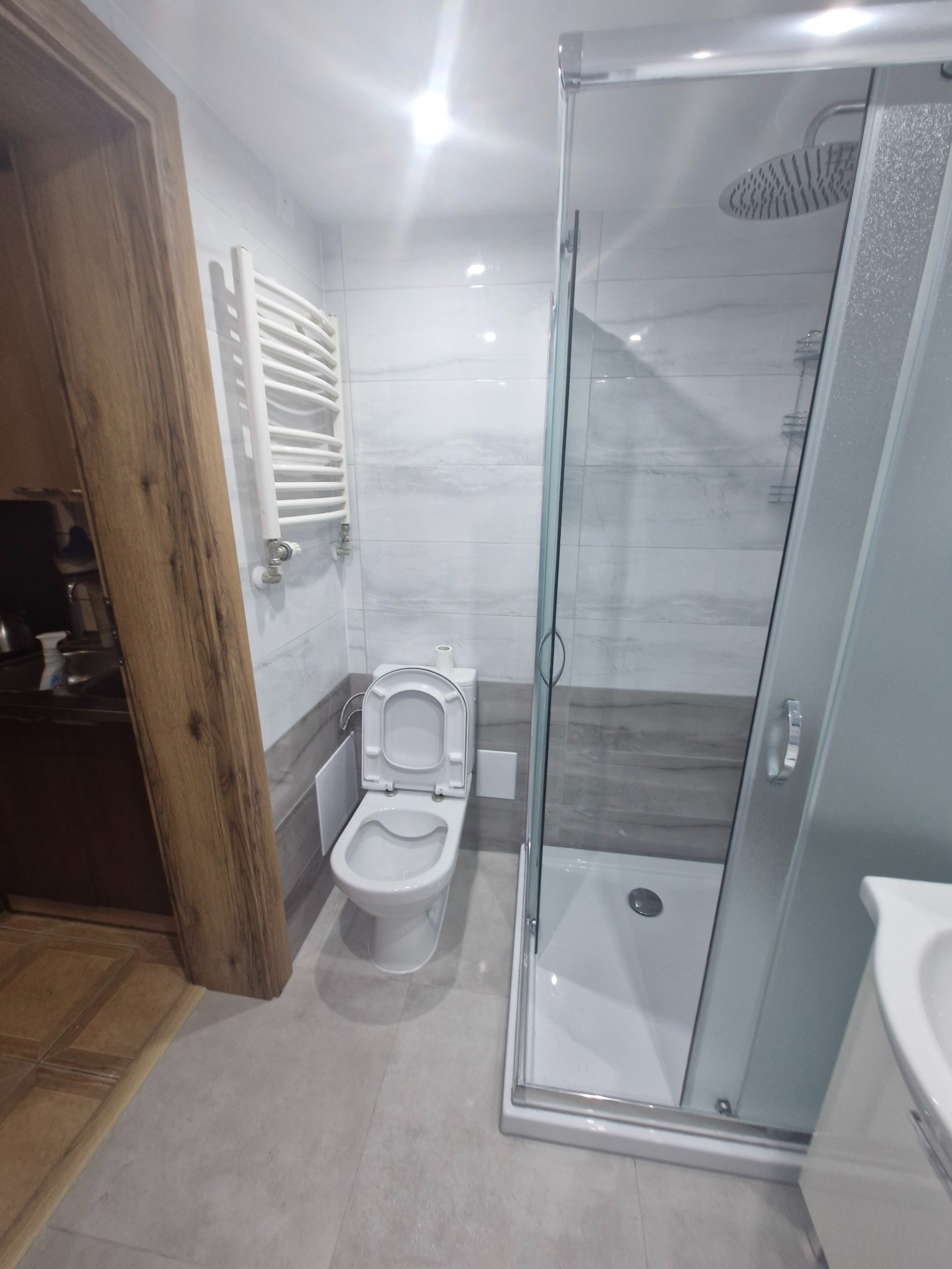 Wykończona łazienka w Działdowie z kabiną prysznicową typu walk-in, podwieszaną toaletą z otwartą klapą i grzejnikiem drabinkowym na ścianie wyłożonej płytkami imitującymi marmur.