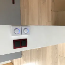 Dwa białe panele dotykowe z niebieskim podświetleniem obok czerwonego włącznika światła na białej ścianie, widok z góry, jasne drewniane panele podłogowe w tle.