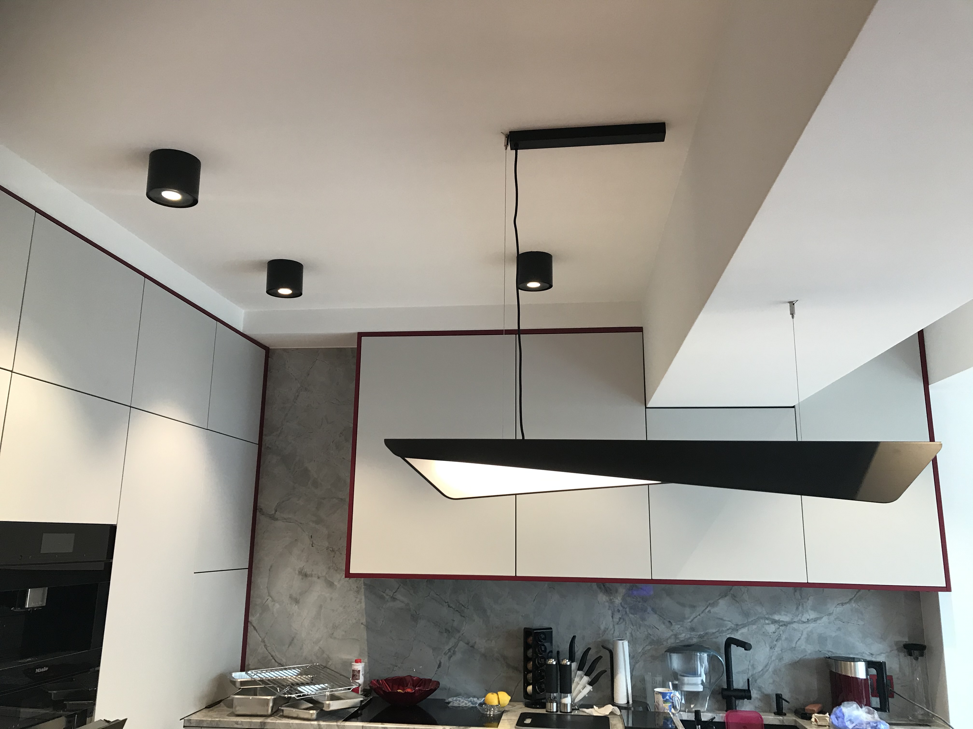 Nowoczesna kuchnia z zamontowanym czarnym oświetleniem sufitowym: podłużna lampa wisząca i trzy okrągłe reflektory punktowe.