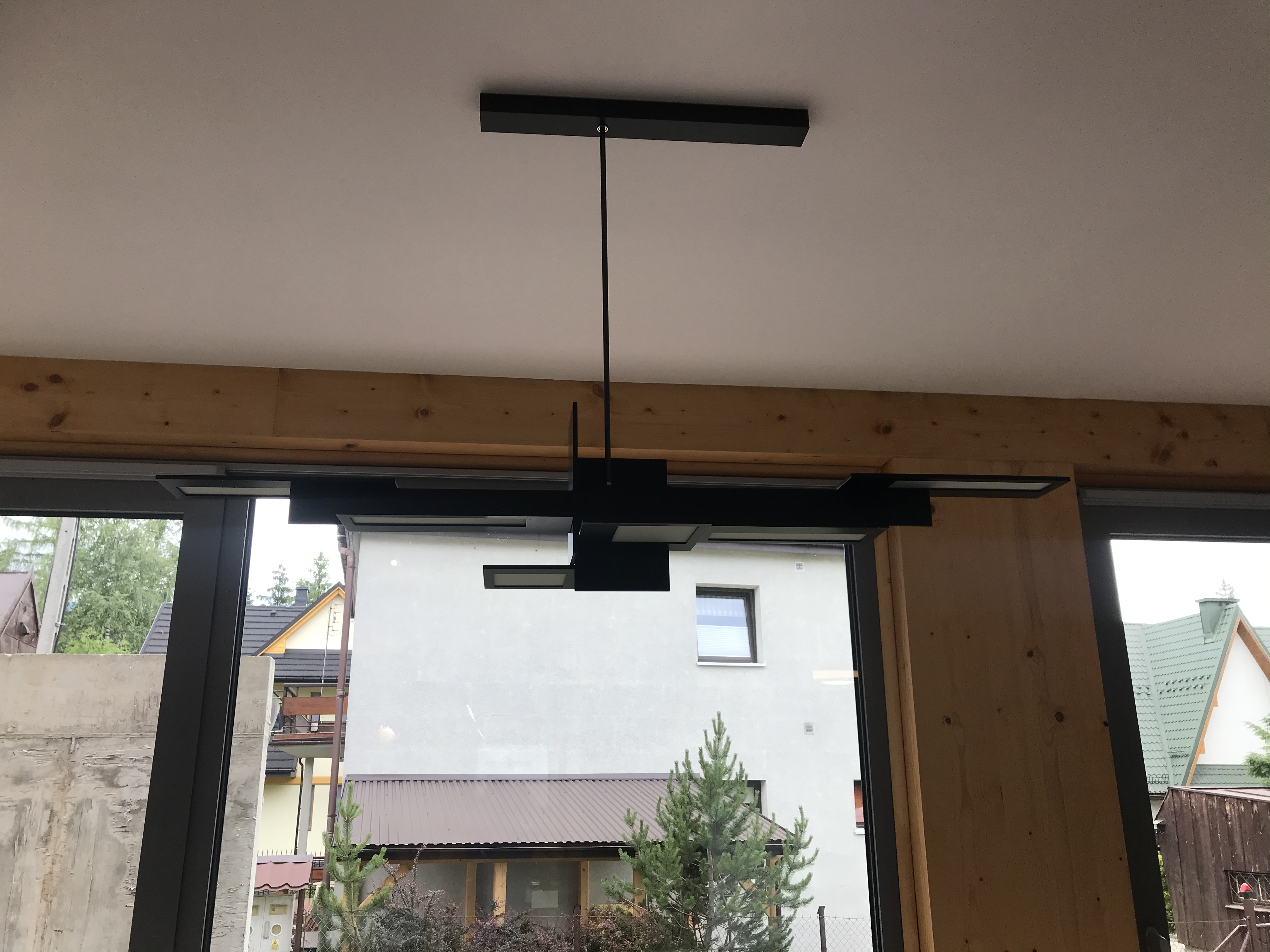 Czarna, geometryczna lampa wisząca zamontowana w nowym domu, widok przez okno na sąsiednie budynki i zieleń.