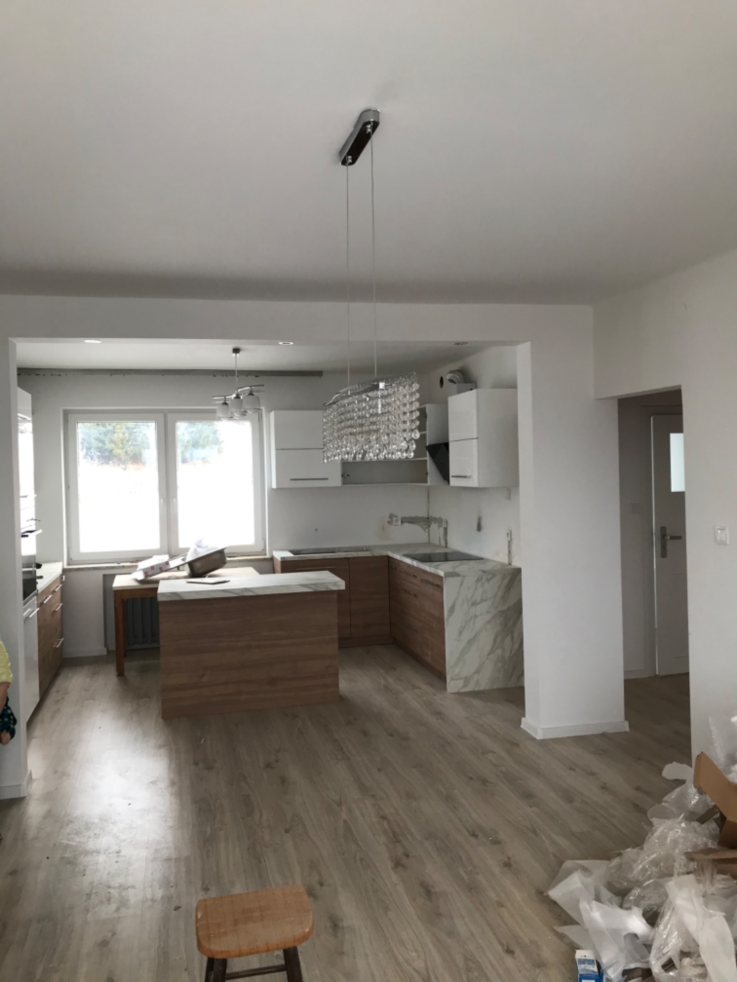 Wnętrze jasnej kuchni w trakcie remontu, widoczna wyspa z drewnianym frontem i marmurowym blatem, kryształowy żyrandol, podłoga panelowa, ślady prac wykończeniowych.