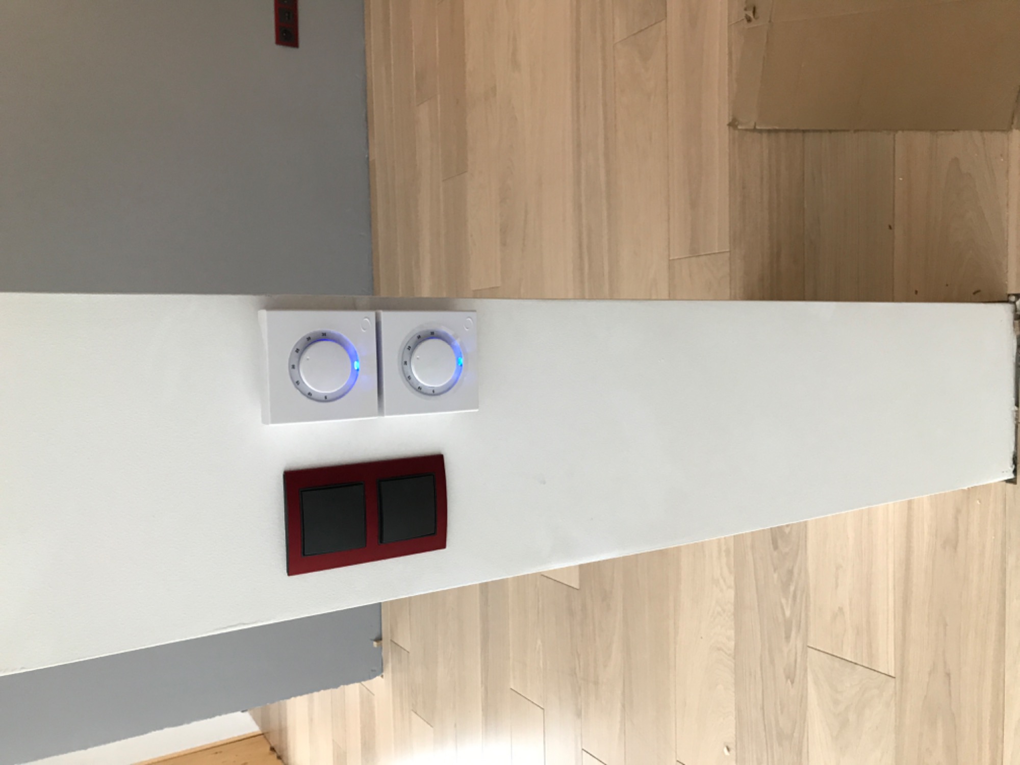 Dwa białe panele dotykowe z niebieskim podświetleniem obok czerwonego włącznika światła na białej ścianie, widok z góry, jasne drewniane panele podłogowe w tle.