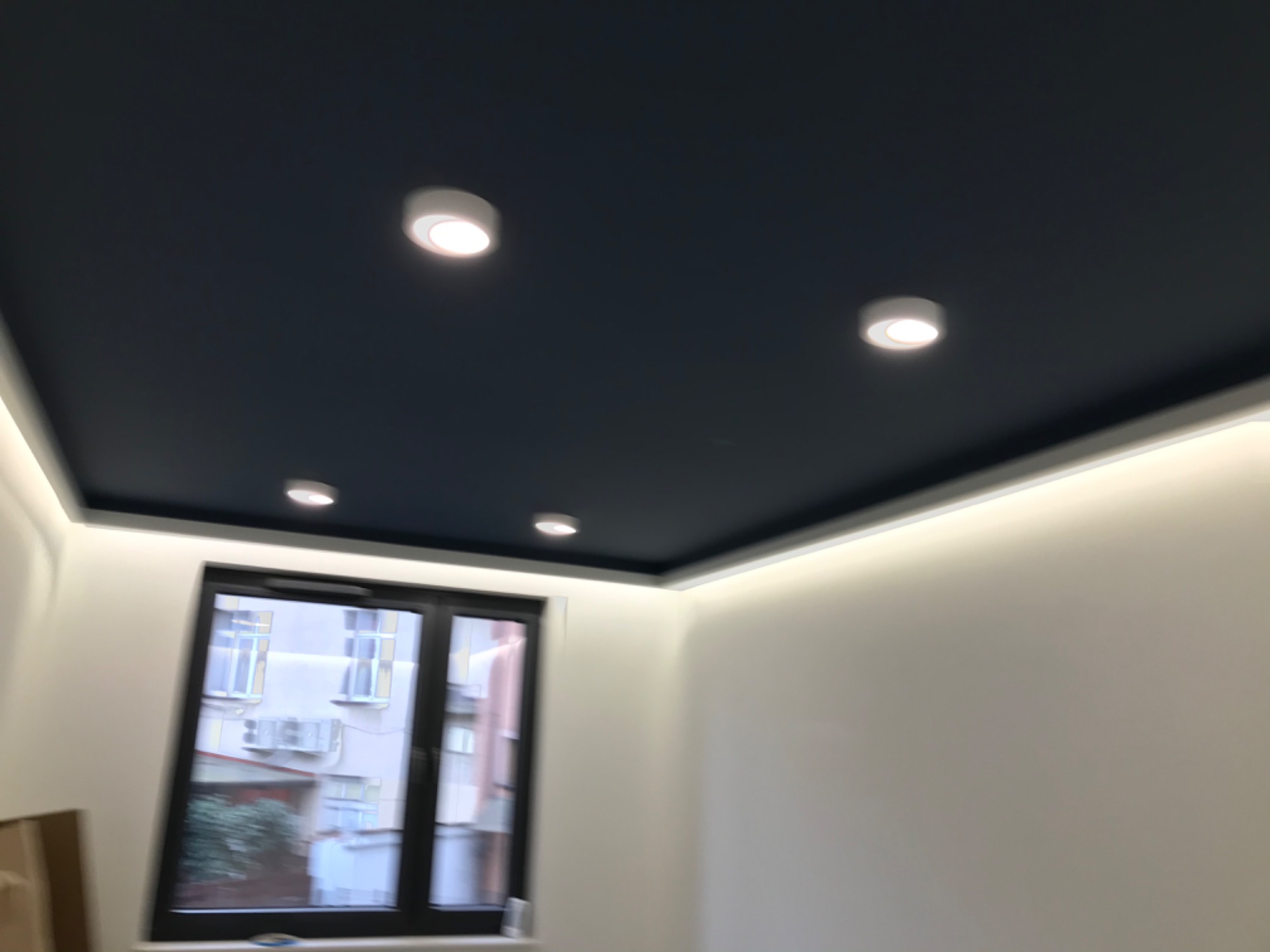 Pomieszczenie z niebieskim sufitem podwieszanym, oświetleniem LED wzdłuż krawędzi i kilkoma okrągłymi lampami punktowymi, widok przez okno na budynek z klimatyzatorami.