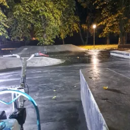 Nocny widok na skatepark z betonową nawierzchnią, oświetlony lampą uliczną, z maszyną do polerowania posadzek z niebieskim wężem na pierwszym planie.