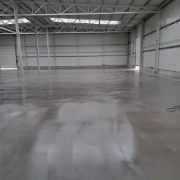 Błyszcząca, szara posadzka przemysłowa w hali magazynowej z metalową konstrukcją dachu i oknami dachowymi. Widok na gładką powierzchnię i elementy konstrukcyjne budynku.