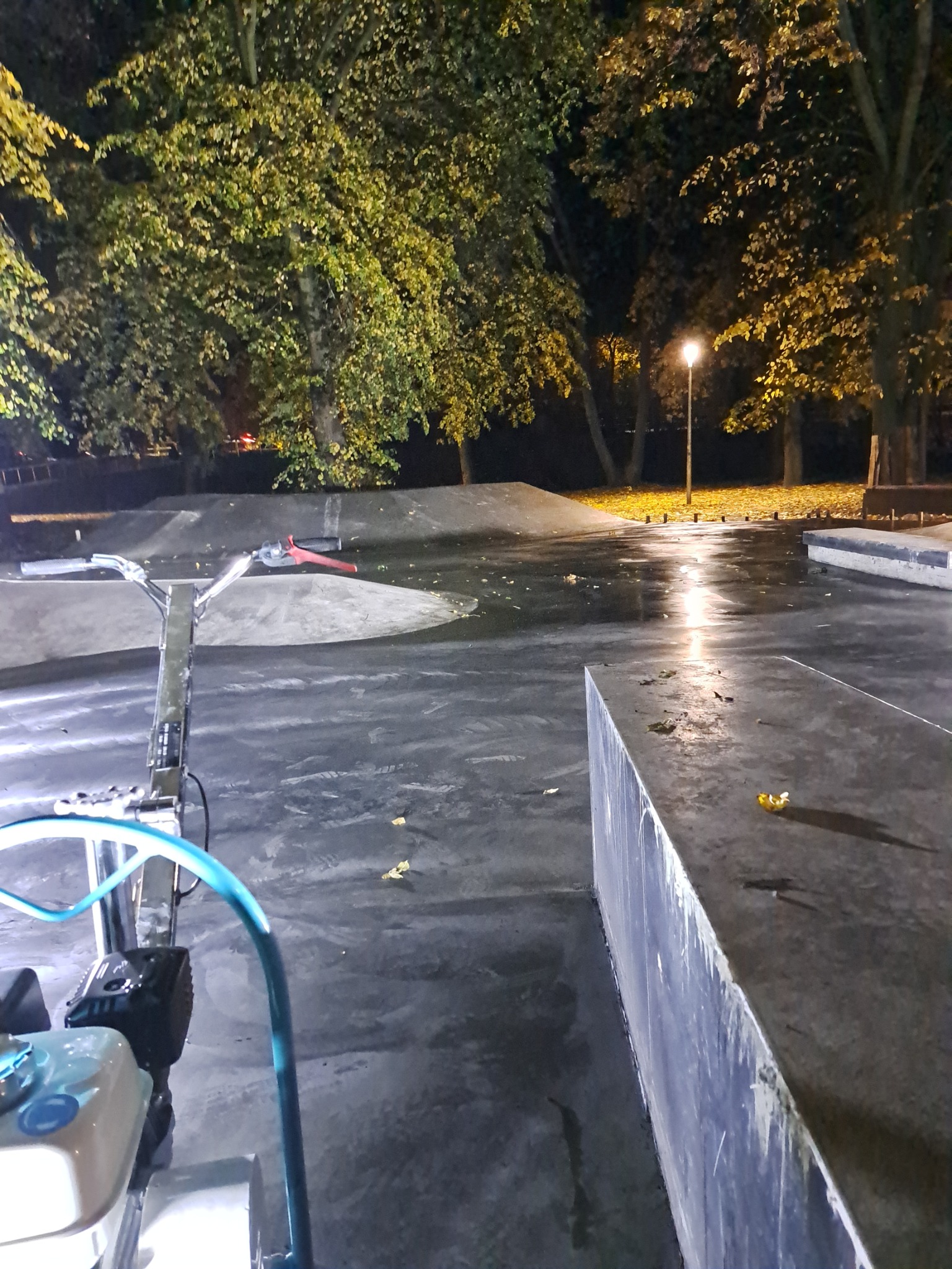 Nocny widok na skatepark z betonową nawierzchnią, oświetlony lampą uliczną, z maszyną do polerowania posadzek z niebieskim wężem na pierwszym planie.