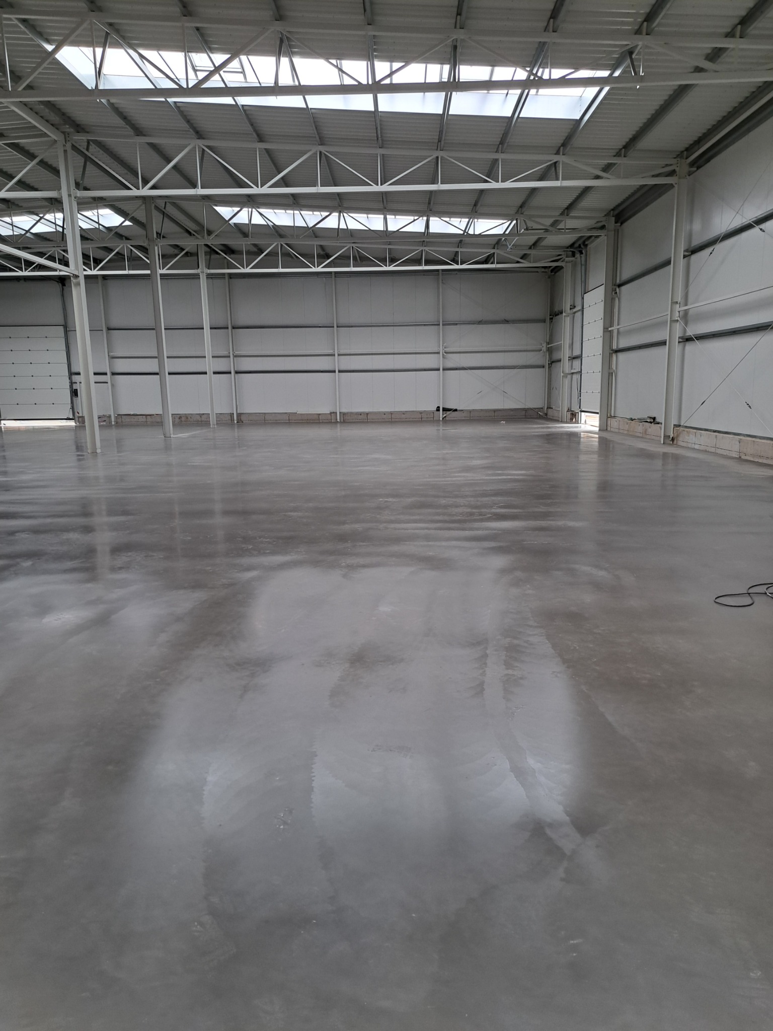 Błyszcząca, szara posadzka przemysłowa w hali magazynowej z metalową konstrukcją dachu i oknami dachowymi. Widok na gładką powierzchnię i elementy konstrukcyjne budynku.