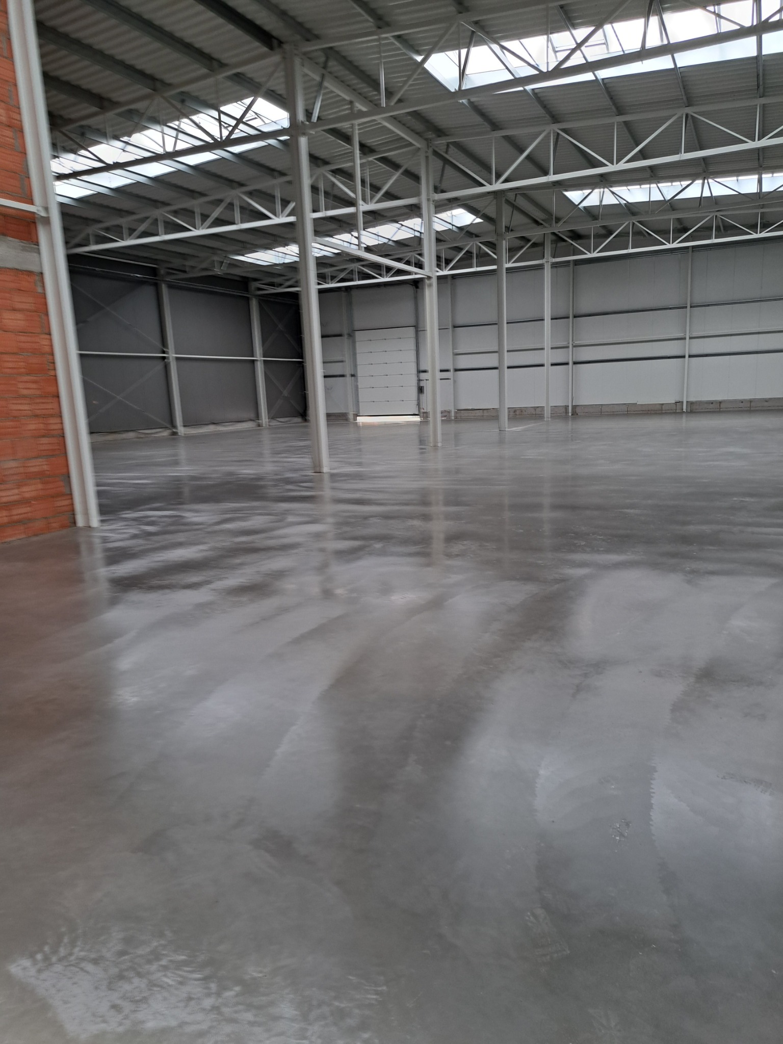 Błyszcząca, szara posadzka przemysłowa w hali magazynowej. Widoczne metalowe słupy konstrukcyjne i fragment ściany z czerwonej cegły. Naturalne oświetlenie z okien dachowych.
