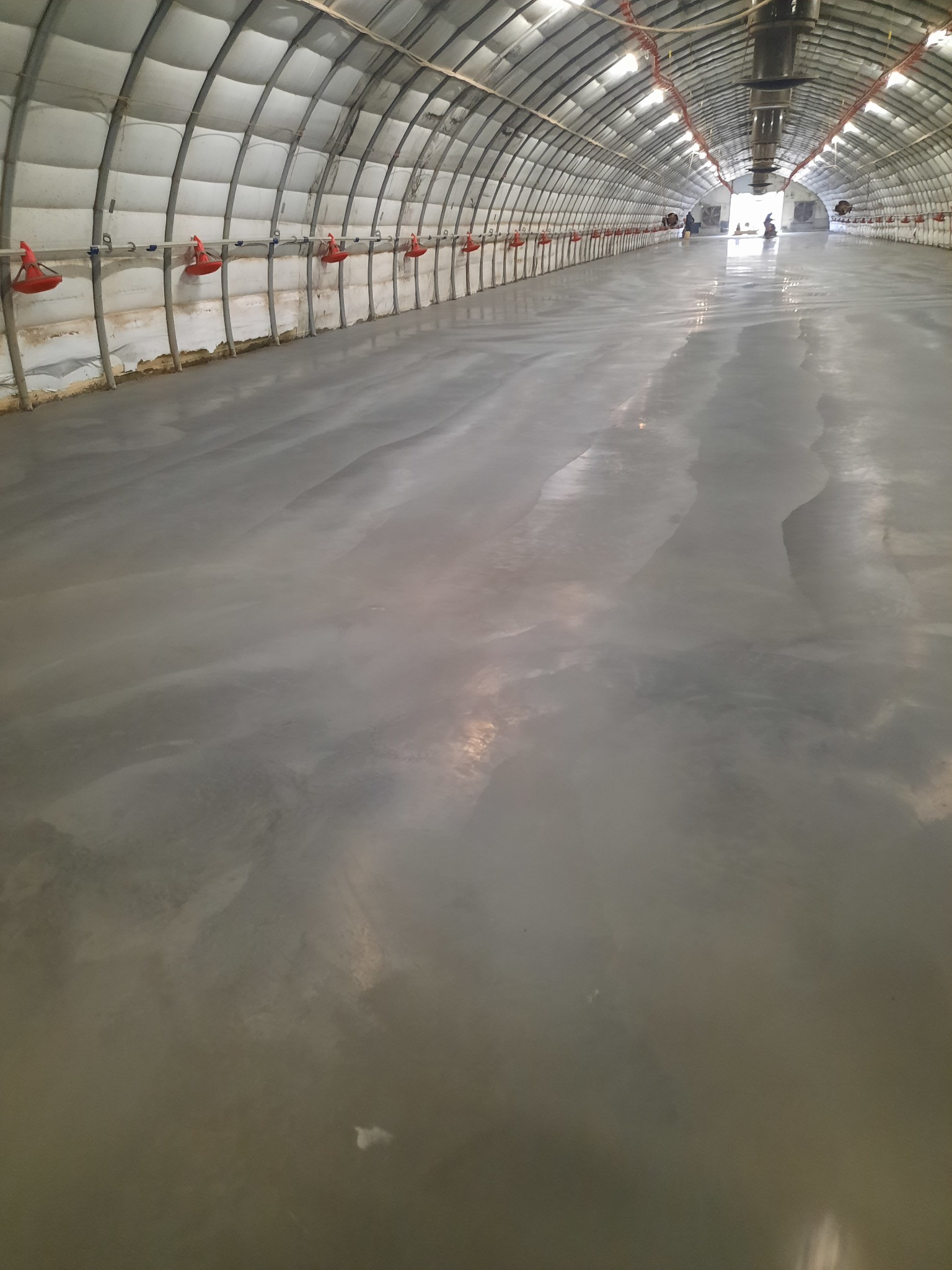 Gładka, szara posadzka przemysłowa w hali z metalową konstrukcją łukową i czerwonymi elementami. Widok w perspektywie, światło odbija się od powierzchni.