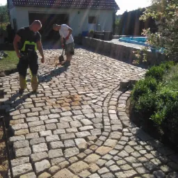 Układanie kostki brukowej z jasnoszarego kamienia na podjeździe, widoczni dwaj pracownicy podczas robót, w tle dom z garażem i basen.