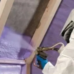 Aplikacja pianki PUR w przestrzeni poddasza z widoczną konstrukcją drewnianą i szczelinami wypełnionymi izolacją, wykonywana przez osobę w kombinezonie ochronnym.