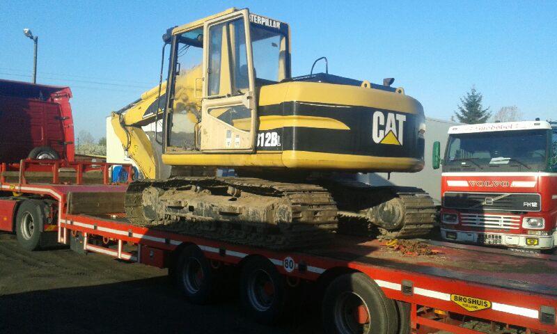 Żółto-czarna koparka Caterpillar 12B na czerwonej platformie transportowej z widocznymi ciężarówkami w tle, ujęcie z boku.