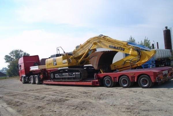 Żółta koparka Komatsu załadowana na czerwoną naczepę transportową, widoczna kabina ciągnika siodłowego i fragmenty innych pojazdów w tle.