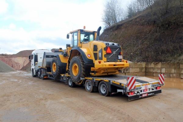 Żółta ładowarka Volvo L150H przewożona na niskopodwoziowej platformie przez białą ciężarówkę MAN, na tle wzgórza i wyrobiska.