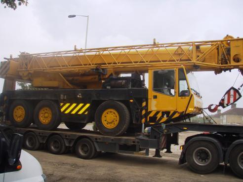 Żółty dźwig samojezdny na niskopodwoziowej naczepie transportowej, widoczne pasy ostrzegawcze i hak dźwigowy.